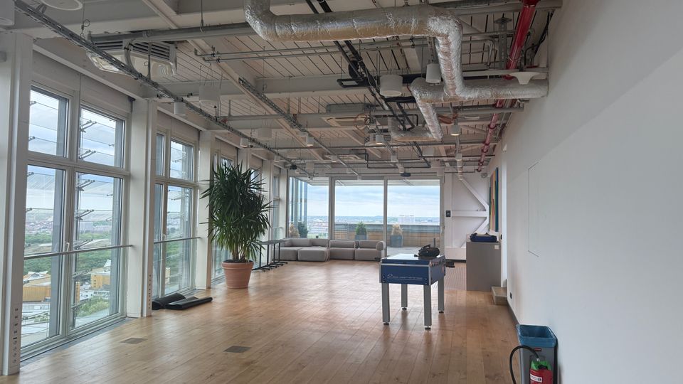 Spacious modern office lounge with floor-to-ceiling windows overlooking a city view, wood plank flooring, and an exposed industrial ceiling with ducts and piping. The open area includes a large potted plant, a gray sectional sofa, and a foosball table near a white wall.