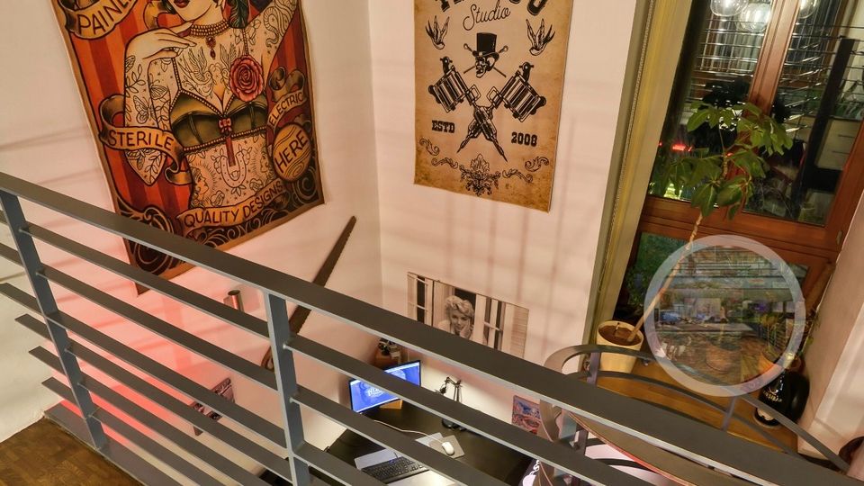 Innenansicht eines Tattoo-Studios mit hoher, weißer Wand und gewölbter Ziegeldecke, gesehen von einer Galerie mit Metallgeländer und Holzboden. An der Wand hängen große Tattoo-Poster, daneben ein hohes Fenster; darunter ist ein Arbeitsplatzbereich mit Schreibtisch und Ausstattung erkennbar.