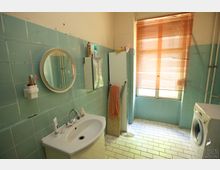 Badezimmer mit hellblauen Fliesen an den Wänden, einem weißen Waschbecken mit rundem Spiegel, einer Waschmaschine, einem Fenster mit Jalousien und einem Schrank. Der Boden ist mit weißen Fliesen ausgelegt.