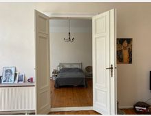 Ein Innenraum mit Doppelflügeltür aus Holz, die in ein Schlafzimmer führt. Im Schlafzimmer sind ein Metallbett mit grauer Bettdecke, ein Hängelüster, eine Korbstuhl-Ecke und Holzdielenboden zu sehen; im Vordergrund links steht eine Kommode mit gerahmten Bildern und daneben eine Wand mit einem Gemälde.