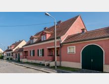 Reihe von niedrigen Wohnhäusern in einer gepflasterten Straße, charakterisiert durch rosafarbene Fassaden, grüne Fensterläden und Satteldächer mit Gauben; im Vordergrund eine Straßenlaterne und ein grünes Scheunentor.