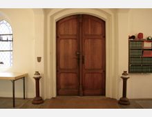 Holztür in einem weißen Innenraum mit rundbogigem Rahmen, flankiert von zwei schmalen Säulenständern. Links ein Tisch und ein gotisches Fenster mit verzierten Bleiglas-Scheiben, rechts ein Bücherregal mit grünen und roten Büchern.