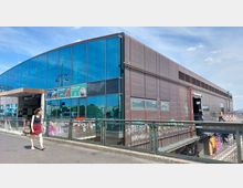 Modernes Bahnhofsgebäude mit brauner Metallfassade und großen blauen Glasfronten, unter anderem mit Werbeschildern beklebt. Im Vordergrund führt ein Gehweg mit Geländer entlang, während im unteren Bereich eine untere Passage mit bunten Graffiti sichtbar ist.