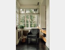 Wohnbereich mit großer Fensterfront, die auf einen bewaldeten Außenbereich blickt. Im Vordergrund stehen ein schwarzer Sessel, ein Korb mit einer Decke und eine Pflanze auf einer Fensterbank; helle Wände und dunkler Holzboden ergänzen das schlichte Interieur.