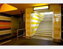 U-Bahn-Station mit einem beleuchteten Treppenaufgang, gelb-weißen Fliesen an den Wänden und mehrfarbigen Wandverkleidungen auf der linken Seite. Ein Geländer führt entlang der Treppe, und der Boden ist dunkel.