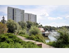 Hohes Wohnhaus im Hintergrund, umgeben von Bäumen, einem Fußweg im Vordergrund und einem Wasserlauf mit angelegten Booten rechts im Bild. Der Himmel ist klar mit wenigen Wolken.
