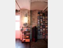 Industriell gestalteter Innenraum mit unverputzten Backsteinwänden, einem Metallregal mit Dekorationsgegenständen wie einem Globus und Körben sowie einem hohen schwarzen Lautsprecher. Rechts im Bild befindet sich ein großer, unordentlich gestapelter Bücher- und DVD-Turm.