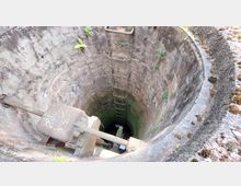 Blick in einen alten, runden Brunnenschacht mit gemauerten Wänden, an denen stellenweise Pflanzen wachsen. Im Inneren sind eine Metallleiter und rostige Mechanikteile sichtbar.