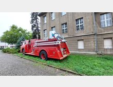 Ein rotes, historisches Feuerwehrfahrzeug mit Schläuchen und Metallaufbauten steht auf einer Rasenfläche neben einem großen, beige verputzten Gebäude mit mehreren Fenstern. Im Hintergrund sind Bäume und ein weiteres Gebäude zu sehen.