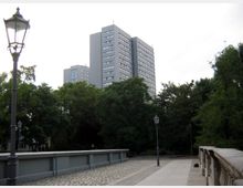 Ein moderner mehrstöckiger Gebäudekomplex im Hintergrund, umgeben von Bäumen, mit einer gepflasterten Brücke und antiken Straßenlaternen im Vordergrund. Der Himmel ist bewölkt, und die Umgebung wirkt parkähnlich.