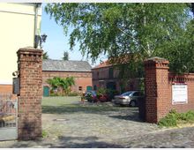 Backstein-Hofanlage mit gepflastertem Innenhof, umgeben von roten Ziegelgebäuden mit grünen Türen und Fenstern. Im Vordergrund ein Eingangstor mit Ziegelpfeilern und einer Beschilderung, sowie ein Baum und parkende Autos im Hof.