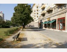 Straßenansicht mit breitem Gehweg, einer Reihe von Bäumen und links angrenzendem Grünstreifen; rechts befinden sich helle Wohngebäude mit Balkonen und Geschäften im Erdgeschoss, darunter eines mit grüner Markise. Im Vordergrund steht eine Holzbank auf dem Gehweg.