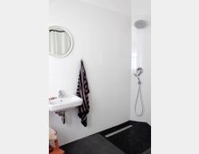 Modernes Badezimmer mit weiß gefliesten Wänden, einem runden Spiegel über einem kleinen Waschbecken, einer aufgehängten gestreiften Handtuch, einer offenen Dusche mit Regenduschkopf und einem dunklen Bodenbelag mit flachem Abfluss.