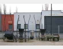 Mehrere Handkarren und Schubkarren stehen in einer Reihe vor einem modernen, zweifarbigen Gebäude mit weißer und roter Fassade. Der Bereich ist von einem Metallzaun umgeben; im Hintergrund sind Fenster, Türen und eine große graue Garagentür zu sehen.