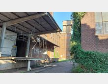 Außenansicht eines verlassenen Industriegebäudes mit rostbraunen Ziegelwänden, einem überdachten Ladebereich mit Metallgerüst und einem hohen Gebäudeturm im Hintergrund. Pflanzen und Efeu überwuchern Teile der Fassade, und der Boden besteht aus Kopfsteinpflaster.