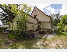 Seitenansicht eines zweigeschossigen Wohnhauses mit hell verklinkerter Fassade und steilem Satteldach, neben einem kleinen gepflasterten Terrassenbereich mit Überdachung und Stufen. Umgeben von Bäumen, Sträuchern und Rasen führt ein schmaler Weg zur Rückseite, daneben steht ein kleiner Schuppen.