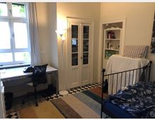 Helles Schlafzimmer mit einem Schreibtisch und Stuhl neben einem Fenster, einem Kleiderschrank mit Glastüren, einem schmiedeeisernen Bett sowie einem offenen Regal. Der Boden ist mit einem schwarz-weißen Fliesenmuster und einem Teppich gestaltet.