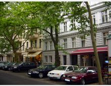 Straßenansicht mit mehrstöckigen Wohnhäusern im Altbaustil, großen Fenstern und dekorativen Fassadenelementen. Vor den Häusern stehen geparkte Autos, während Bäume mit dichtem Blattwerk den Gehweg und die Straße säumen.