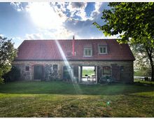 Steinhaus mit roten Ziegeldach in ländlicher Umgebung, umgeben von Bäumen und einer Wiese. Zwei große Türen geben den Blick durch das Haus auf eine grüne Landschaft und einen See im Hintergrund frei.