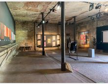 Industrieller Ausstellungsraum mit unverputzten Backsteinwänden, einer offenen Decke mit sichtbaren Rohren und Beleuchtung, sowie mehreren Kunstwerken und Fotografien an den Wänden. Im Vordergrund stehen schwarze Metallpfeiler, eine Kleiderstange mit Kleidung und ein kleiner Podest, während der Raum eine rustikale, renovierte Atmosphäre vermittelt.