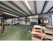 Große Lagerhalle mit Metallstützen und einer niedrigen Decke aus Stahlträgern. Im Raum befinden sich Regale mit gelagerten Materialien, Stapel von Holzpaletten und Arbeitsflächen, der Boden ist grünlich gefärbt.