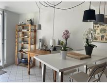 Helle Küche mit weißem Fliesboden, einem langen weißen Esstisch mit Klappstühlen und dekorativen Orchideen in Töpfen. Im Hintergrund ein Holzregal mit Geschirr und eine Wand mit botanischem Poster.