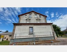 Ehemaliges, renovierungsbedürftiges Gebäude mit der Aufschrift „Lehrlingswohnheim“ an der Fassade. Vor dem Gebäude steht ein Bauzaun, und im Hintergrund sind Wiesen sowie Nebengebäude sichtbar, unter einem blauen Himmel mit Wolken.