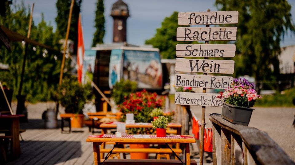 Holzterrasse eines Außenrestaurants mit Picknicktischen und rot-weiß karierten Tischdecken, dekoriert mit Blumen und Schweizer Flaggen. Im Vordergrund steht ein handgemaltes Holzschild mit Speise- und Getränkeangeboten; im Hintergrund sind Bäume und ein Turm zu sehen.