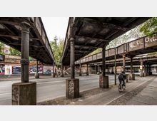 Straßenansicht mit einer alten Eisenbahnüberführung aus Metall, gestützt von verzierten Säulen. Darunter verläuft eine mehrspurige Straße mit Gehweg, an deren Rand Plakatwerbungen und Graffiti auf einer Backsteinmauer sichtbar sind.