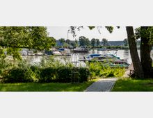 Eine Uferlandschaft mit kleinen Booten, die im Wasser an einem Steg festgemacht sind. Im Vordergrund befinden sich ein gepflasterter Weg, grüne Büsche, hohe Gräser und Bäume, die teilweise den Blick auf die Boote verdecken.