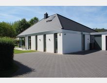 Modernes Einfamilienhaus mit weißer Fassade und flachem Walmdach, großen Glasfenstern und einer angrenzenden Terrasse. Die Umgebung umfasst einen gepflasterten Hof, Rasenflächen und Hecken, umgeben von Bäumen im Hintergrund.