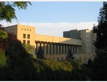 Großes, modernes Gebäude mit hellen Fassaden und großzügigen Säulen entlang einer verglasten Front, umgeben von dichter Vegetation und Bäumen. Der Himmel ist klar mit leichtem Sonnenlicht, das die Architektur beleuchtet.