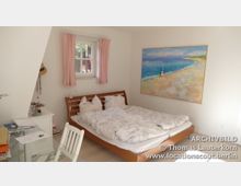 Helles Schlafzimmer mit einem Holzbett, weißem Bettzeug und einem großen Gemälde an der Wand, das eine Strandlandschaft zeigt. Auf der linken Seite sind ein Schreibtisch mit Papieren und ein kleines Fenster mit rosa Vorhängen sichtbar.