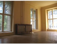 Heller Innenraum mit gelben Wänden, großen Doppelfenstern mit Sprossen und einem Heizkörper unter einem der Fenster. Der Raum hat einen einfachen Fußboden und wirkt leer.