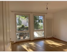 Helles, leeres Zimmer mit Holzboden und großen Fenstern, die auf einen grünen Außenbereich blicken. An der rechten Wand befindet sich ein weißer Heizkörper, und von der Decke hängt eine Glühbirne ohne Lampenschirm.