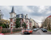 Straßenansicht in einem Wohngebiet mit historischer Villa im Vordergrund, die rote Backstein- und weiße Stuckelemente sowie einen Turm mit spitzem Dach aufweist. Am Straßenrand parken Autos, darunter ein roter Oldtimer, und im Hintergrund sind weitere Wohnhäuser und Bäume zu sehen.