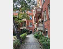 Backsteingebäude mit verzierten Balkonen und weißen Fenstern, umgeben von einem kleinen gepflasterten Weg mit Grünflächen und Sträuchern; ein schräg wachsender Baum ragt in das Bild hinein.