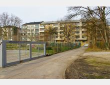 Mehrstöckiges Wohngebäude mit teils verputzter Fassade im Hintergrund, eingerahmt von kahlen Bäumen. Im Vordergrund ein metallisches Schiebetor und ein umzäunter Bereich, sowie eine asphaltierte Zufahrtsstraße.