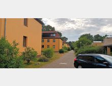 Städtische Wohnsiedlung mit gelb gestrichenen Mehrfamilienhäusern entlang eines gepflasterten Weges, umgeben von kleinen Büschen und Bäumen; im Vordergrund parkt ein schwarzes Fahrzeug. Der Himmel ist bewölkt.