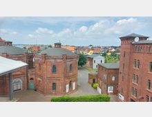 Backsteinbauten einer ehemaligen Industrieanlage mit runden und eckigen Türmen, umgeben von einem gepflasterten Innenhof; im Hintergrund eine Kleinstadt mit Wohnhäusern und Bäumen unter bewölktem Himmel.