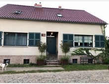 Haus mit cremefarbener Fassade, rotem Ziegeldach und grünen Fensterläden. Die Vorderseite des Hauses zeigt zwei Fenster, eine mittige Haustür, und ein kleines, gepflastertes Vorgartenstück mit einem Baum, einer Bank und einem niedrigen Zaun aus Klinkersteinen. Ein Hund läuft auf dem Kopfsteinpflaster davor.