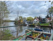 Flusslandschaft mit Wohnhäusern am Ufer, darunter ein modernes Gebäude und traditionelle Einfamilienhäuser mit Garten. Im Vordergrund ein mit Graffiti bemaltes Geländer, im Fluss fährt ein kleines Motorboot.