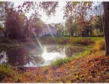 Ein ruhiger Park mit einem kleinen Teich im Vordergrund, umgeben von Herbstlaub und Bäumen. Im Hintergrund steht ein helles, zweistöckiges Gebäude, das von Sonnenlicht bestrahlt wird.