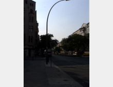 Straßenszene in einer Stadt mit mehrstöckigen Wohn- und Geschäftshäusern am Straßenrand. Ein geschwungener Straßenlaternenmast und vereinzelte Bäume säumen die breite, asphaltierte Straße.