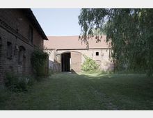 Backsteinhof mit offenen Scheunentoren, umgeben von Rasen und Bäumen. Das Gebäude besitzt ein rotes Ziegeldach sowie kleine Fenster und hölzerne Türläden.