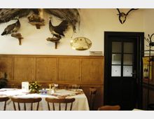 Innenraum eines traditionellen Gasthauses mit Holztäfelung an den Wänden, einem dunkel gerahmten Glastürbereich und ausgestopften Pfauen auf Wandregalen. Ein gedeckter Tisch mit Blumenarrangement und Reservierungsschildern befindet sich im Vordergrund.