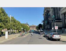 Straßenszene mit Blick auf einen historischen Platz, der von Bäumen gesäumt ist. Rechts im Vordergrund die Fassade des Berliner Doms, im Hintergrund das Alte Museum mit seiner klassizistischen Architektur. Auf der Straße sind Autos, E-Roller und Fußgänger zu sehen.