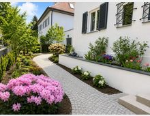 Geschwungener Kopfsteinpflasterweg durch einen gepflegten Vorgarten mit Blumenbeeten und Sträuchern neben einem weißen Wohnhaus mit schwarzen Fensterläden und Fenstergittern. Im Hintergrund stehen weitere helle Häuser mit Satteldach, umgeben von Bäumen.