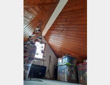 Ein Dachbodenraum mit einer schrägen Holzdecke, einer kleinen Fensteröffnung mit Tageslichteinfall und mehreren gestapelten Kunststoffboxen zur Lagerung. Links steht eine minimalistische Metallstruktur in Form eines Weihnachtsbaums, dekoriert mit roten Kugeln.