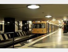 U-Bahn-Station mit einem gelben Zug der Berliner U-Bahn-Linie U7, Ziel "Rudow". Der Bahnsteig hat eine helle, geflieste Oberfläche mit schwarzen und weißen Details, die Wände sind teilweise mit schwarzen, glänzenden Säulen verkleidet, und die Decke ist mit runden Deckenleuchten ausgestattet.